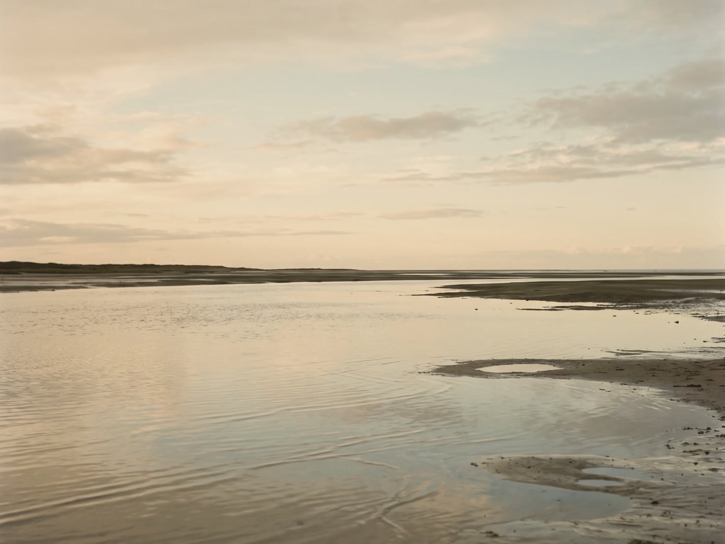 Stil ondiep water bij laagwater met zandplaten, een lage duinrand aan de horizon en een lichte lucht met zachte wolken
