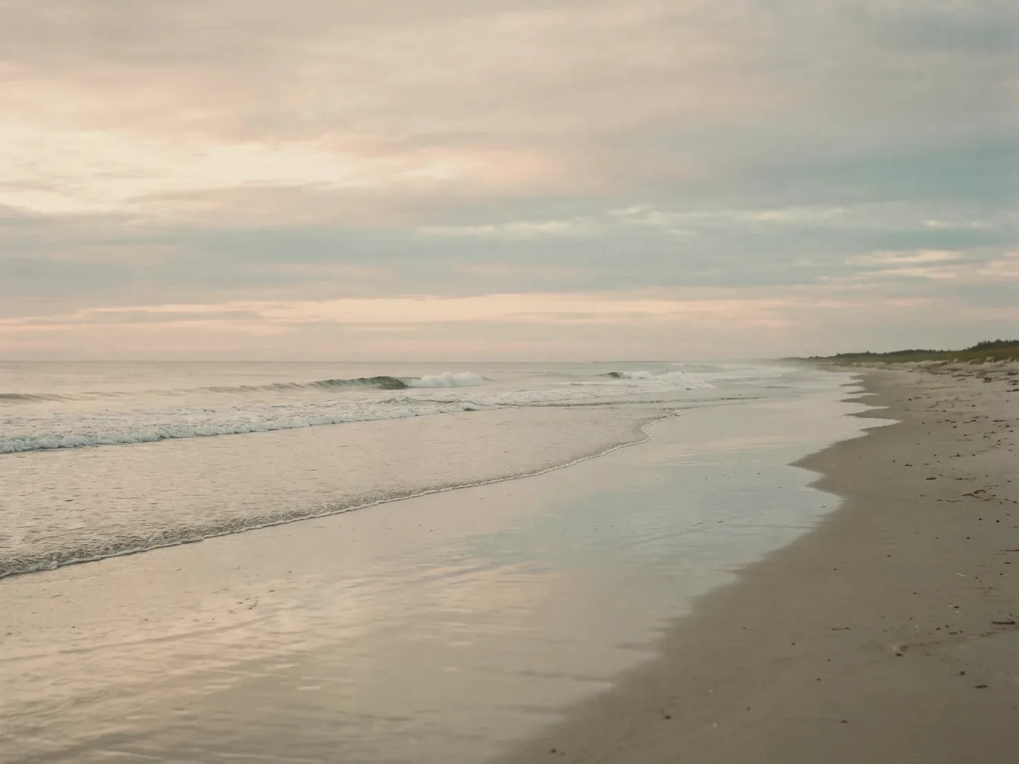 Een rustige zee bij zonsopkomst, met een vlakke golf die over glad nat zand uitloopt onder een zachtroze en grijze lucht, en rechts een leeg strand met begroeide duinen.