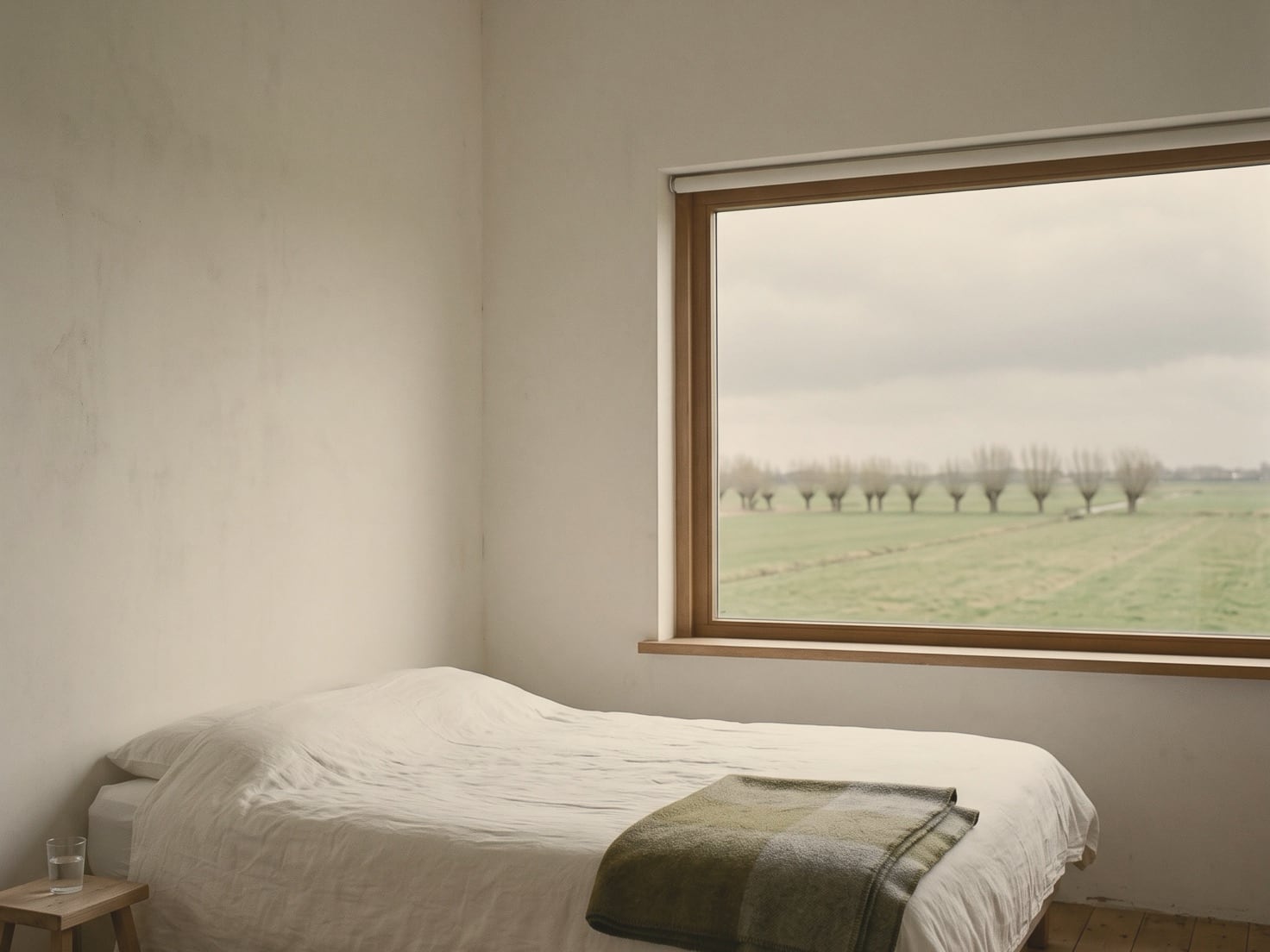 Een eenvoudige kamer met een opgemaakt bed in wit linnen en een groene wollen deken, een krukje met een glas water en een groot raam dat uitkijkt op vlakke groene weilanden met een rij knotwilgen onder een grijze lucht