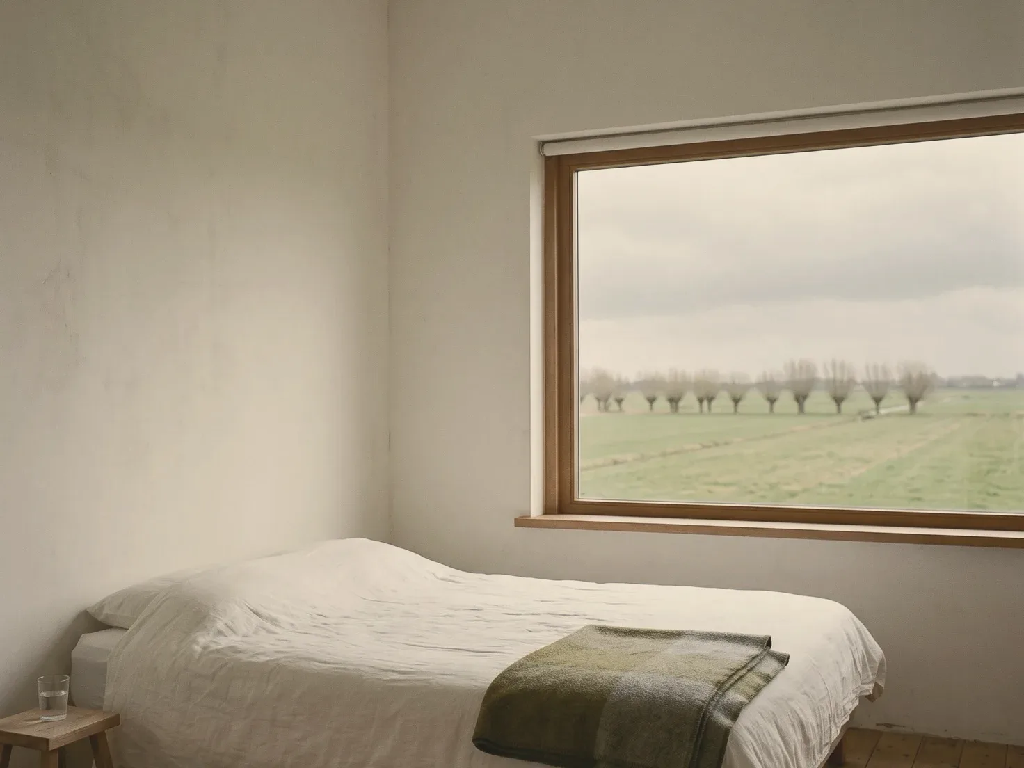 Een eenvoudige kamer met een opgemaakt bed in wit linnen en een groene wollen deken, een krukje met een glas water en een groot raam dat uitkijkt op vlakke groene weilanden met een rij knotwilgen onder een grijze lucht