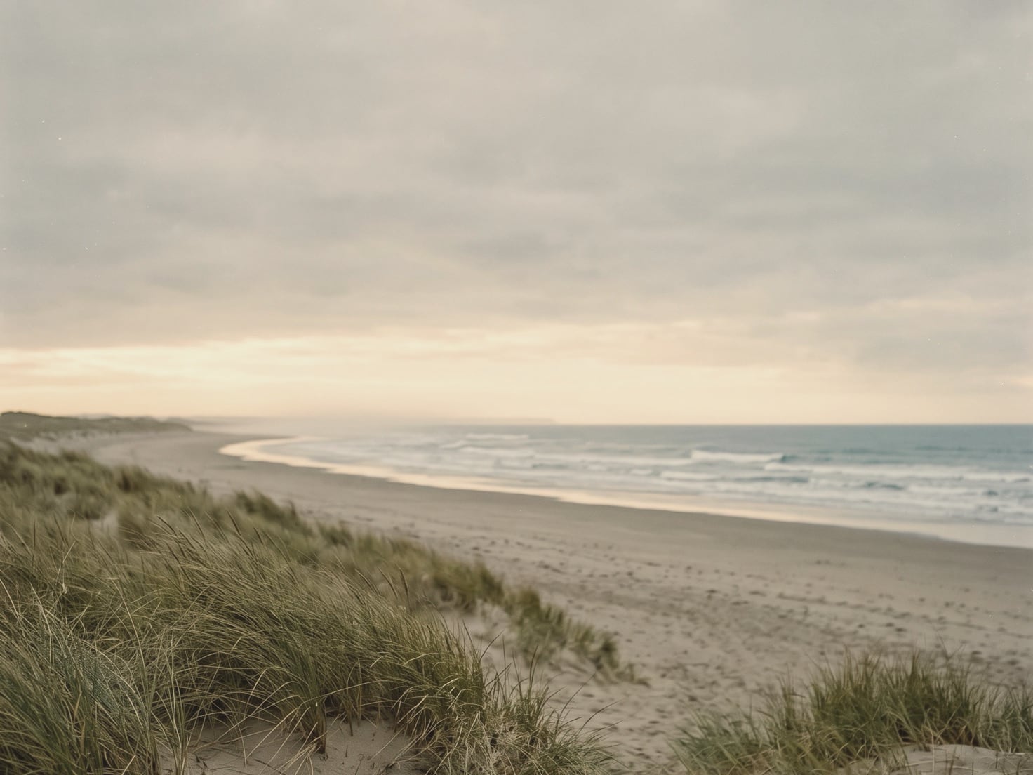 Een breed, leeg strand onder een grijze bewolkte lucht met een lichte strook aan de horizon, lage golven die aanrollen en helmgras op een duin op de voorgrond