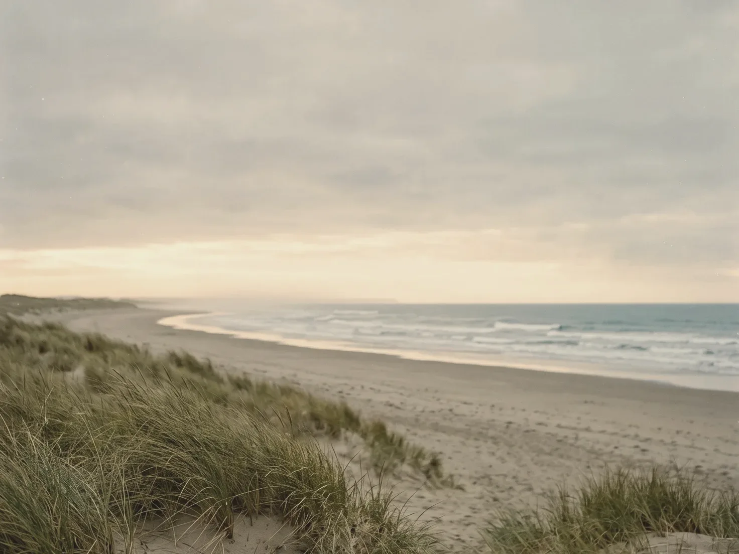 Een breed, leeg strand onder een grijze bewolkte lucht met een lichte strook aan de horizon, lage golven die aanrollen en helmgras op een duin op de voorgrond